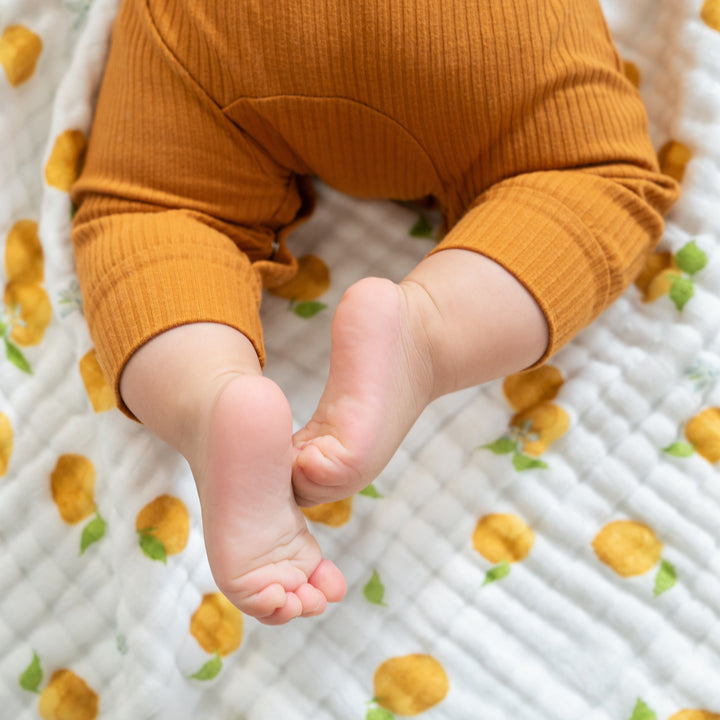 A baby wearing orange ribbed pants relaxes on an aden + anais Organic Baby Essentials Bundle Dream Blanket, showcasing a white and yellow lemon pattern, with their legs and bare feet in view.