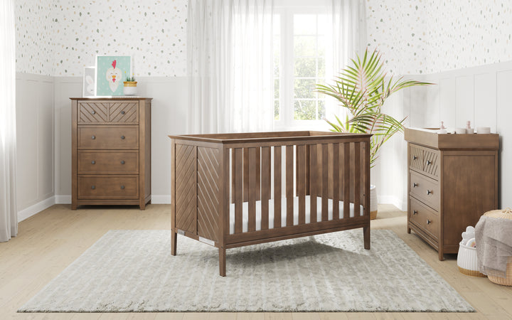 A bright nursery features a wooden crib on a light rug, the Child Craft Atwood 3-Drawer Dresser in Cocoa Bean, matching chest, a plant by the window, soft curtains, and patterned wallpaper with white paneled walls.