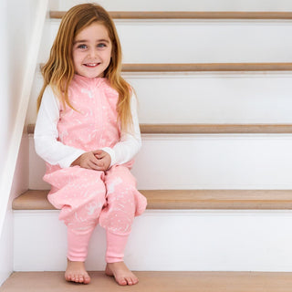 A young girl with long light brown hair, wearing a pink patterned onesie over a white top, sits barefoot on wooden stairs, smiling at the camera.