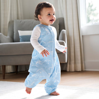 A smiling baby in a blue and white cloud-patterned onesie walks barefoot indoors on a light rug, with a chair and window in the background.