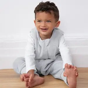 A young child with curly hair, wearing a light gray outfit and sitting barefoot on a wooden floor, smiles while reaching for their toes.