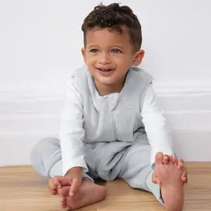 A young child with curly hair, wearing a light gray outfit and sitting barefoot on a wooden floor, smiles while reaching for their toes.