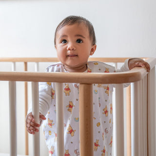 A baby wearing a white outfit with cartoon prints stands and holds onto the wooden rails of a crib, looking up with a curious expression.