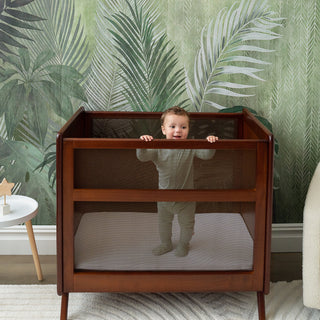 A baby stands holding onto the sides of a wooden crib with mesh panels, set against a green leafy wallpaper. A small white table with wooden toys is nearby.