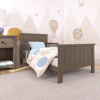 A wooden toddler bed with a colorful dotted comforter and blue pillow, placed on a textured rug in a childs room with hot air balloon and star-themed wall decorations.