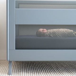 A baby wrapped in a beige swaddle lies on a dark mattress inside a modern gray crib with mesh sides, on a textured light-colored carpet.