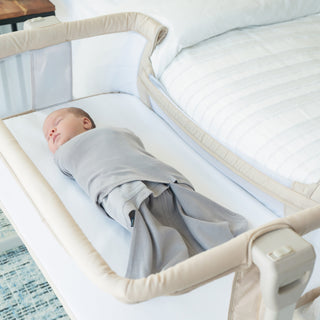A baby wrapped in a gray swaddle sleeps peacefully in a beige bedside bassinet next to a neatly made white bed.