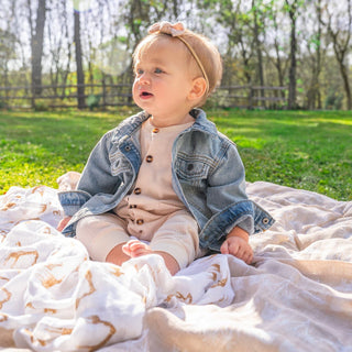 A baby wearing a denim jacket and headband sits on a blanket in a sunny park. Trees and grass are in the background.