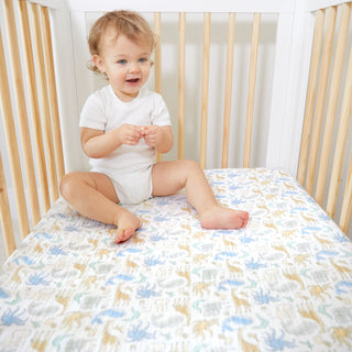 A smiling toddler in a white onesie sits in a crib with light wooden bars, on a crib sheet decorated with colorful dinosaur illustrations.