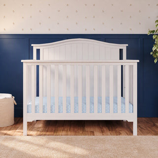 Matte white Child Craft Hampton Arch Top 4-in-1 Convertible Crib with vertical slats and a light blue polka dot mattress sheet.