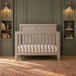 Child Craft Kieran 4-in-1 Convertible Crib in Crescent Gray, featuring a paneled headboard and slatted sides, set in a room with green paneled walls and built-in shelving.