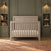 Child Craft Kieran 4-in-1 Convertible Crib in Crescent Gray, featuring a paneled headboard and slatted sides, set in a room with green paneled walls and built-in shelving.