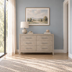 A gray wooden Kieran 6-Drawer Double Dresser, Crescent Gray, with long metal pulls, topped with a table lamp, a small bowl, books, and a carved animal figure.