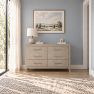A gray wooden Kieran 6-Drawer Double Dresser, Crescent Gray, with long metal pulls, topped with a table lamp, a small bowl, books, and a carved animal figure.