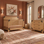 The Child Craft Lucas 2-Piece Nursery Set, featuring a wooden crib and a three-drawer dresser with dark metal corner accents, is placed in a room with mauve paneled walls.