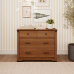 A Child Craft Redmond 3 Drawer Dresser in Coach Cherry featuring matte black hardware, topped with a storage basket, books, and a potted plant.