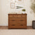 A Child Craft Redmond 3 Drawer Dresser in Coach Cherry featuring matte black hardware, topped with a storage basket, books, and a potted plant.