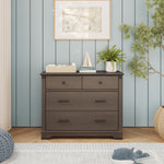 A Dapper Gray Child Craft Redmond 3 Drawer Dresser with one top drawer and two wide lower drawers, topped with a woven basket, a toy sailboat, and a potted plant.