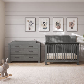 Child Craft Denman 4-in-1 Convertible Crib in Midnight Gray and matching six-drawer dresser with a changing top, positioned against a shiplap wall with four framed botanical prints.