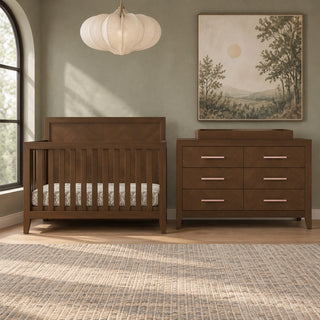 A dark wood Kieran 4-in-1 Convertible Crib in Toasted Chestnut and a matching six-drawer dresser with a changing topper in a nursery.