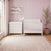 A matte white Child Craft Atwood 4-in-1 Convertible Crib and a matching three-drawer changing table sit in a nursery with pink striped wallpaper and a patterned rug.