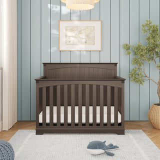 A dark wood Child Craft Redmond 4 Piece Nursery Set convertible crib with slats stands against a light blue paneled wall as the centerpiece of the room.
