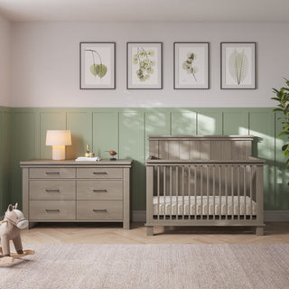 A Child Craft Denman Double Dresser, Crescent Gray, and a matching slatted crib sit side by side against a sage green paneled wall.