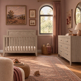 A Child Craft Kieran 6-Drawer Double Dresser in Crescent Gray with six drawers and metal pulls, situated in a nursery next to a matching wooden crib.