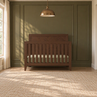 A Child Craft Kieran 4-in-1 Convertible Crib in toasted chestnut with a chevron pattern on the headboard, positioned against an olive green paneled wall.