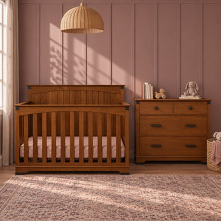 A Child Craft crib and matching Redmond 3 Drawer Dresser in Coach Cherry sit on a patterned rug in a room with paneled walls.