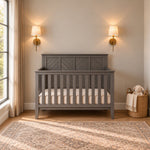 Child Craft Atwood 4-in-1 Convertible Crib in Lunar Gray, featuring a diagonal slat design on the headboard, positioned against a wall between two wall-mounted lamps.