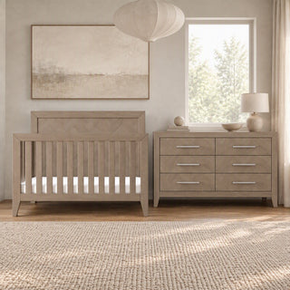 A light gray Kieran 6-Drawer Double Dresser with six horizontal silver pulls stands next to a matching crib in a nursery.