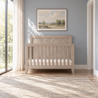 A Child Craft Kieran 4-in-1 Convertible Crib in Crescent Gray with a chevron pattern on the headboard stands on a woven rug in a room.