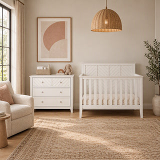 A white Child Craft Atwood 3-Drawer Dresser, Matte White with a changing table topper and a matching white slatted crib in a nursery, with a woven pendant light hanging above.