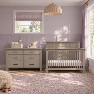 A light-gray Child Craft Denman Double Dresser in Crescent Gray with six drawers stands next to a matching crib in a room with light purple walls.
