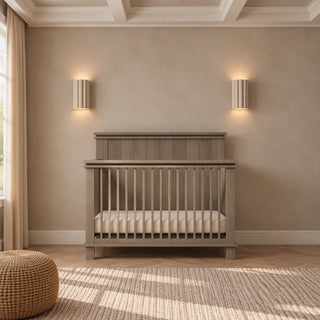 A gray Child Craft Denman 4-in-1 Convertible Crib made of pine wood, featuring slatted sides and a mattress, positioned against a wall with two sconces.