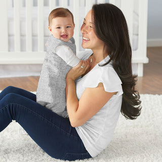 A smiling woman sits on a white rug, cradling a happy baby in a gray HALO Wearable Blanket 100% Cotton TOG 0.5. The bright, airy room with a white crib highlights safe sleeps peace. She wears a white top and dark jeans, enjoying this serene moment.