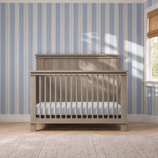 Child Craft Denman 4-in-1 Convertible Crib in Crescent Gray with slatted sides set against a blue and white striped wallpapered wall.