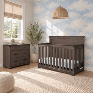 A dark gray Child Craft Redmond wooden crib and matching Redmond 3 Drawer Dresser, Dapper Gray in a nursery with cloud-patterned wallpaper.