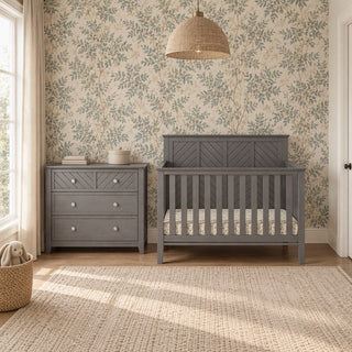 A gray Child Craft Atwood 3-Drawer Dresser, Lunar Gray sits next to a matching crib with a patterned mattress in a nursery.