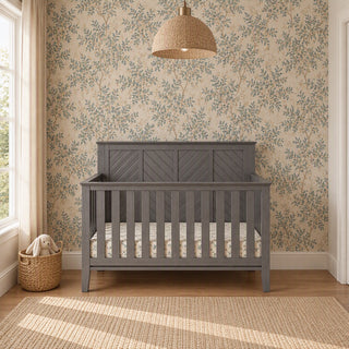 Child Craft Atwood 4-in-1 Convertible Crib in Lunar Gray, featuring a slat design and a patterned mattress against a floral wallpapered wall.