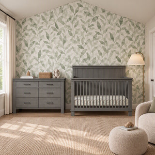 A gray wooden Child Craft Denman Double Dresser, Midnight Gray with six drawers sits next to a matching crib in a room with floral wallpaper and a textured rug.