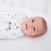 A baby lies on a white surface, wrapped in a HALO Swaddle 100% Cotton TOG 1.5 with colorful triangle patterns, smiling gently and looking relaxed as the adjustable wings promote healthy hip development.