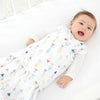A baby lies in a crib, wearing a HALO Swaddle 100% Cotton TOG 1.5 with colorful triangle patterns. The swaddles adjustable wings support healthy hip development. The happy baby looks upwards, resting on a white sheet.