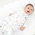 A baby lies in a crib, wearing a HALO Swaddle 100% Cotton TOG 1.5 with colorful triangle patterns. The swaddles adjustable wings support healthy hip development. The happy baby looks upwards, resting on a white sheet.