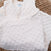 A soft, white HALO Swaddle Plushy Dot Velboa TOG 3.0 with adjustable wings lies on a woven brown blanket. This sleepsack has a zipper and round embroidered patch, promoting healthy hip development for your little one.