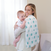 A smiling woman holds a laughing baby over her shoulder with an aden + anais boutique 100% cotton muslin swaddle featuring green dinosaurs. Its breathable cotton ensures excellent absorbency. The room has light beige walls and sheer white curtains.