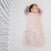 A baby sleeps peacefully on a white mattress, wrapped in a HALO Swaddle 100% Cotton TOG 1.5 with soft pink floral patterns for healthy hip development, while the cribs white slats stand against the striped rug partially covering the floor.