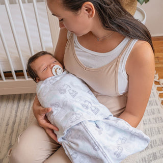 A woman cradles a sleeping baby in a HALO Swaddle Micro-Fleece TOG 3.0, ensuring healthy hip development with its adjustable wings. They are in a cozy room with a crib and rug, creating an atmosphere of comfort and warmth.