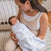 A woman cradles a sleeping baby in a HALO Swaddle Micro-Fleece TOG 3.0, ensuring healthy hip development with its adjustable wings. They are in a cozy room with a crib and rug, creating an atmosphere of comfort and warmth.